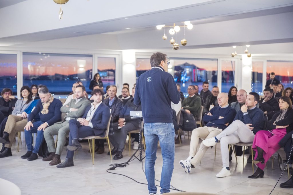 Relatore in piedi che parla a una platea numerosa in una sala con vetrate